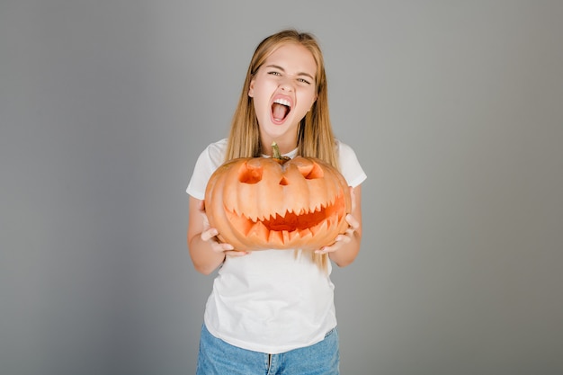 Spielerisches lustiges blondes Mädchen mit dem Halloween-Steckfassung O-Laternenkürbis getrennt über Grau