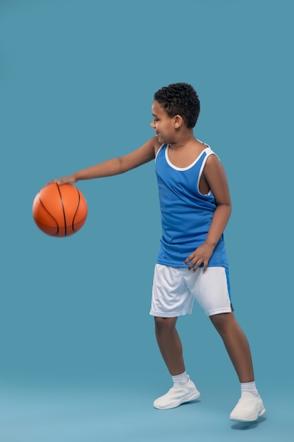 Spielzeit. Dunkelhäutiger Junge in Sporthemd und Shorts, der sich mit Ball im Basketball auf blauem Hintergrund im Studio bewegt