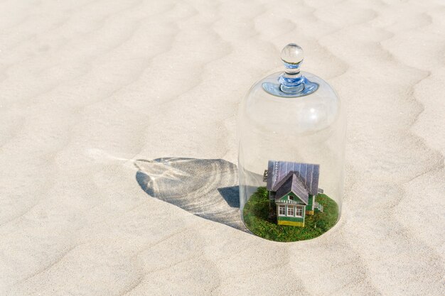 Spielzeug-Kartonhaus mit grünem Rasen, geschützt durch eine Glaskuppelglocke inmitten einer leblosen Sandwüste