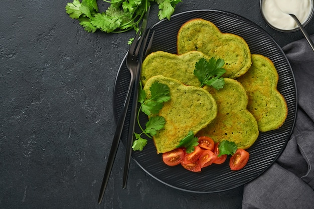 Spinat-Avocado-Pfannkuchen in Herzform mit griechischer Joghurtsauce und Kirschtomaten auf einem schwarzen Teller auf dunklem Hintergrund. Frühstückskonzept. Draufsicht mit Kopienraum.