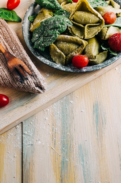 Spinat-Tortellini auf einem alten Holztisch, umgeben von frischen Zutaten: Tomaten, Mehl, Spinat, Eier. Veganes Essen. Copyspace