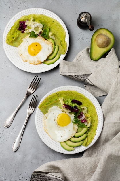 Spinatgrüne Crepes (Pfannkuchen) mit Spiegelei, Avocado und Salatmischungen auf Keramikplatte auf grauer Betonoberfläche. Selektiver Fokus. Draufsicht. Copt Raum.