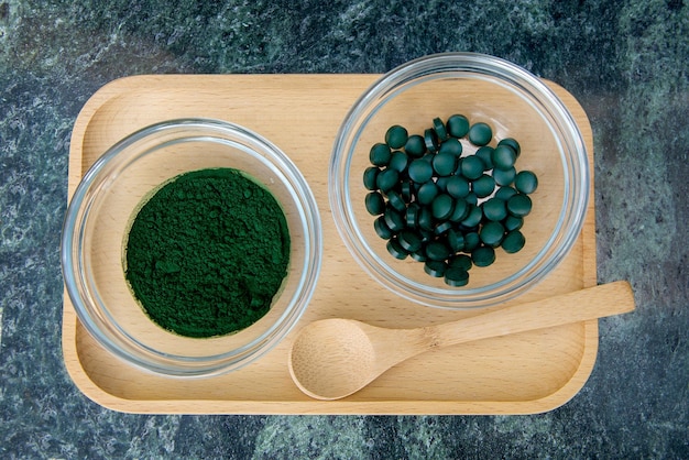 Spirulina-Pulver und Pillen in Glasschalen auf einem Holztablett mit einem Holzlöffel auf grünem Hintergrund