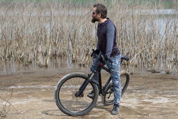 Sport brutaler bärtiger Kerl auf einem modernen Mountainbike. Ein Radfahrer an einem einsamen Ort am See.