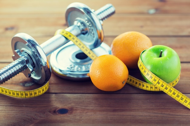 Sport-, Fitness-, Ernährungs- und Objektkonzept - Nahaufnahme von Hantel und grünem Apfel mit Orangen, die mit Maßband auf Holztisch umwickelt sind