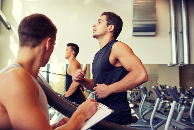 Foto sport, fitness, lifestyle, technologie und personenkonzept - männer mit personal trainer trainieren auf laufband im fitnessstudio