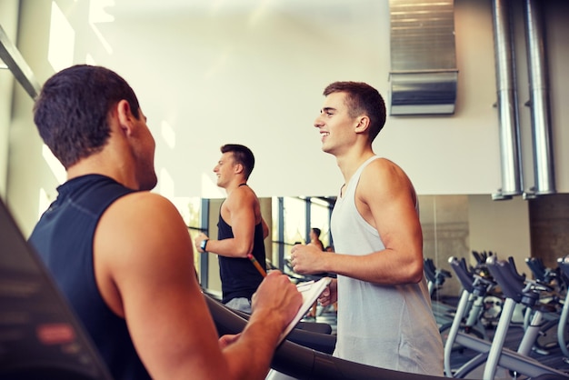 Foto sport, fitness, lifestyle, technologie und personenkonzept - männer mit personal trainer trainieren auf laufband im fitnessstudio