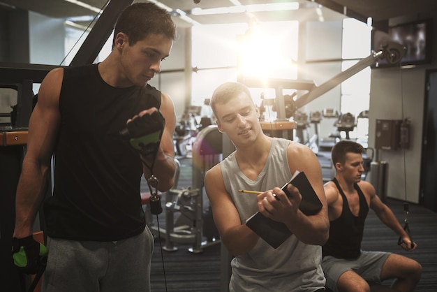 Sport-, Fitness-, Lifestyle- und People-Konzept - Männer mit Klemmbrett, die sich Notizen machen und auf Fitnessgeräten trainieren