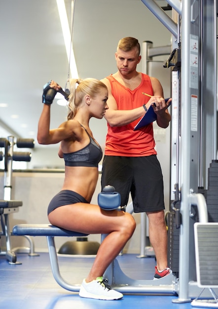 Sport-, Fitness-, Teamwork- und People-Konzept - junge Frau und Personal Trainer, die Muskeln auf einem Fitnessgerät spielen lassen