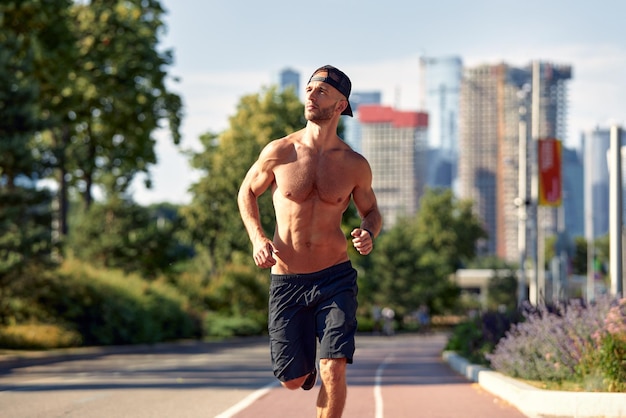 Sport in der Stadt Sportlicher Mann, der an einem schönen Sommertag im Stadtpark ohne Hemd für Fitness joggt