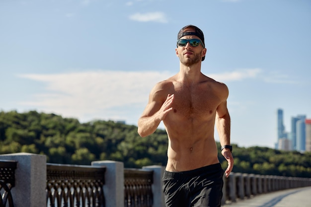 Sport in der Stadt Sportlicher Mann, der an einem schönen Sommertag im Stadtpark ohne Hemd für Fitness joggt