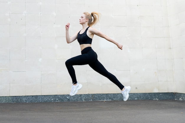 Sport- und Fitnesskonzept Junge Erwachsene kaukasische Sportlerin springen hoch im Freien graue Schwanzwand Hintergrund Sportliche Frau springen Übungen außerhalb gesunder Lebensstil Motivation