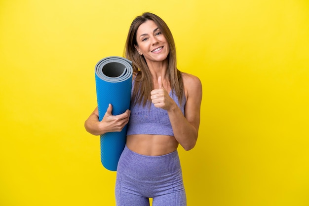 Sportfrau, die zu Yoga-Kursen geht, während sie eine Matte hält, die einen nicht gelben Hintergrund isoliert und eine Daumen-nach-oben-Geste gibt