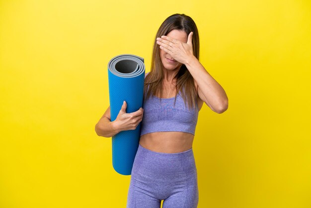 Sportfrau, die zu Yoga-Kursen geht, während sie eine Matte hält, isoliert, nicht gelber Hintergrund, der die Augen mit den Händen bedeckt, will nichts sehen