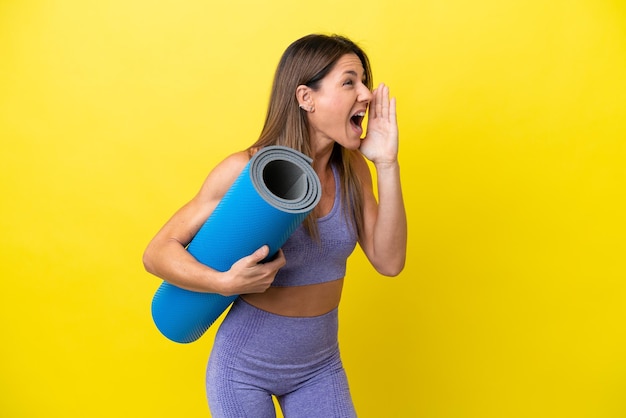 Sportfrau, die zu Yoga-Kursen geht, während sie eine Matte hält, isoliert, nicht gelber Hintergrund, schreit mit weit geöffnetem Mund zur Seite