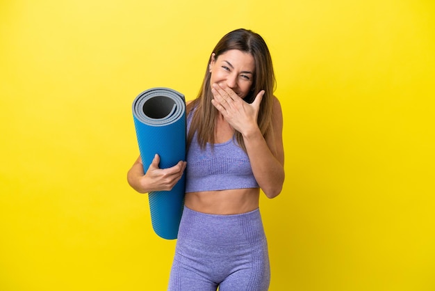Sportfrau, die zu Yogakursen geht, während sie eine Matte hält, isoliert nicht gelber Hintergrund, glücklich und lächelnd, den Mund mit der Hand bedeckend