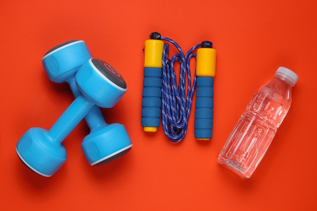Sportkonzept im Flat-Lay-Stil. Kurzhanteln, Springseil, Flasche Wasser. Sportausrüstung auf orange Hintergrund. Draufsicht