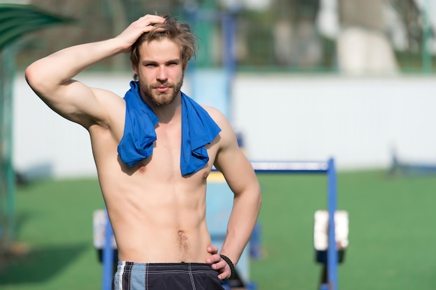 Sportler berühren Haare mit blauem T-Shirt am Hals. Bärtiger Mann mit fitten Rumpfmuskeln im Stadion. Athlet mit sexy Bauch und Brust im sonnigen Freien. Training und Training. Sport oder Fitness und Körperpflege.