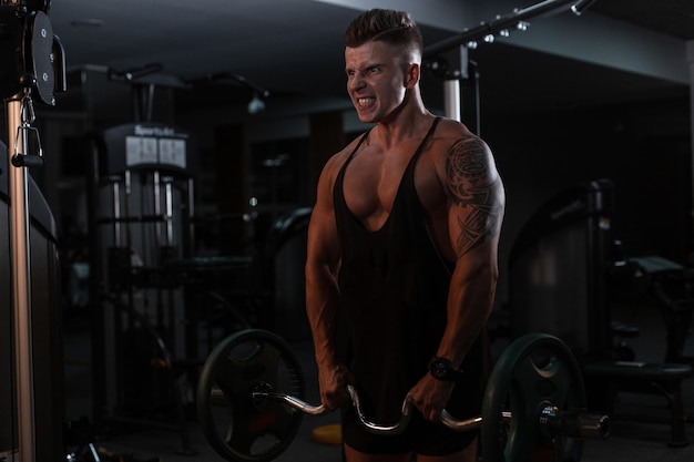 Sportler Bodybuilder mit einem muskulösen Körper in einem schwarzen T-Shirt pumpt ausdrucksvoll seine Muskeln und macht eine Übung im Fitnessstudio