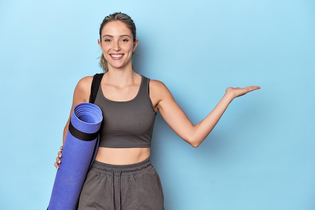 Sportler mit einer Matte im blauen Studio, der einen Kopierraum auf einer Handfläche zeigt und eine andere Hand an der Taille hält