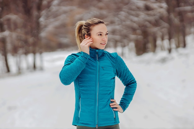 Sportlerin, die an einem verschneiten Wintertag in der Natur mit der Hand auf der Hüfte steht und Kopfhörer aufsetzt. Winterfitness, gesundes Leben, Musik, Spaß