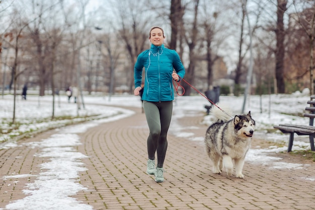 Sportlerin, die bei Schneewetter zusammen mit ihrem Hund im Park joggt. Winterfitness, Haustiere, Freundschaft