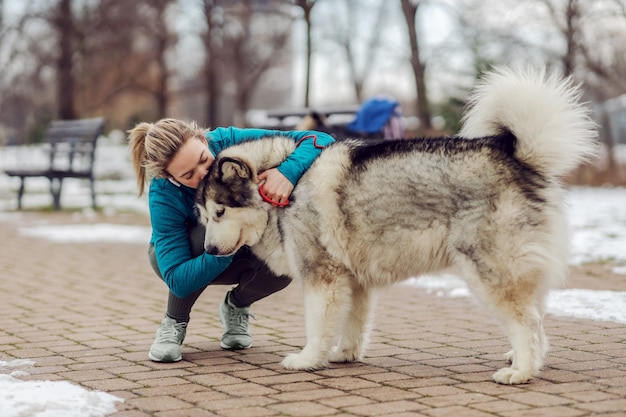 Sportlerin, die ihren Hund küsst und ihn umarmt, während sie bei Schneewetter im Park kauert. Haustiere, Erholung, Wochenendaktivitäten