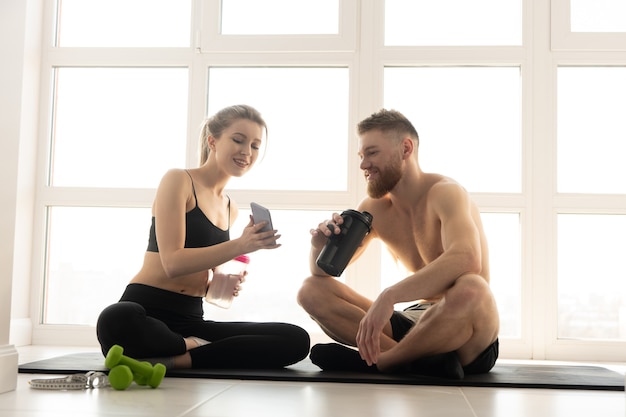 Sportlerin, die Sportler etwas auf dem Handy zeigt. Leute auf Fitnessmatte. Blonde Frau trägt Sportkleidung. Mann mit nacktem Oberkörper. Konzept der sportlichen Aktivität zu Hause. Innenraum der geräumigen sonnigen Wohnung