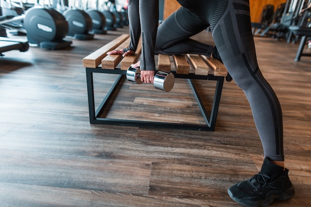 Sportlerin in schwarzer Kleidung mit Hanteln, die auf der Holzbank im Fitnessstudio trainieren