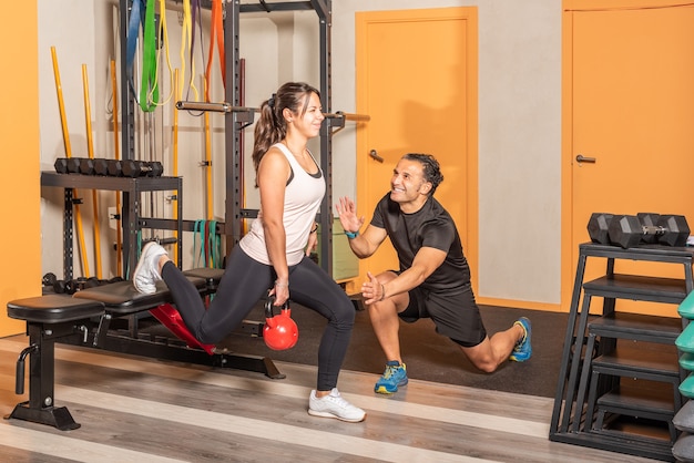 Sportlerin in Sportbekleidung macht Ausfallschritte mit einer Kettlebell in der Hand, wobei ein Bein mit Hilfe des Trainers im Fitnessstudio auf einer Bank ruht. Konzept der Übung mit Geräten im Fitnessstudio.