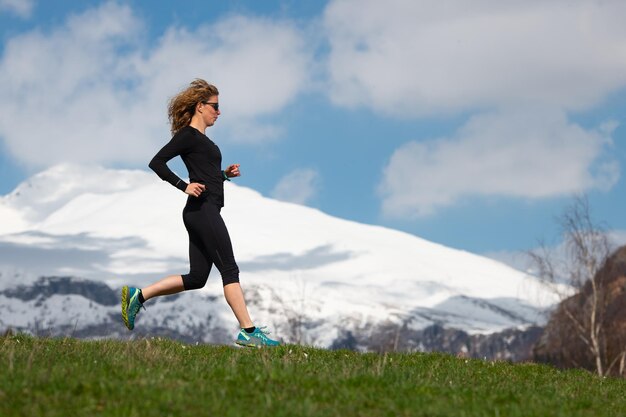 Sportlerin läuft in Bergen auf Frühlingswiese mit Schneegebirgshintergrund