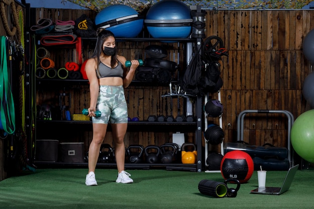 Sportlerin mit Gewichten, die online mit einem Computer unterrichten, der eine Maske für die neue Normalität trägt