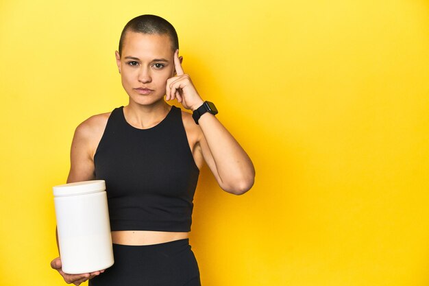 Sportlerin mit Protein-Shake, gelber Studio-Hintergrund, der mit dem Finger auf die Tempel zeigt