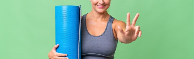 Sportlerin mittleren Alters, die zu Yoga-Kursen geht, während sie eine Matte über isoliertem Hintergrund hält, glücklich und drei mit den Fingern zählt