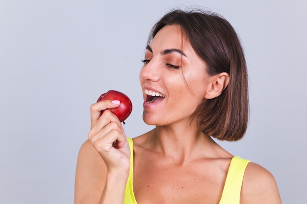 Sportlerin steht auf grauer Wand, zufrieden mit den Ergebnissen von Fitnesstraining und Diät, hält Waage, trägt Top und Leggings, hält Apfel holds