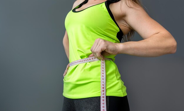 Sportlich fitte Frau, die mit Maßband misst und die Taille nach einer Diät misst, die über grauem Hintergrund isoliert ist. Diätetik Gewichtsverlust Fitness-Größenmessung