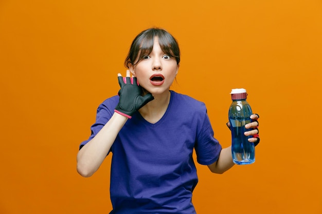 Sportlich schöne Frau in Sportbekleidung in Handschuhen mit einer Flasche Wasser, die in die Kamera blickt und erstaunt und überrascht über orangefarbenem Hintergrund steht