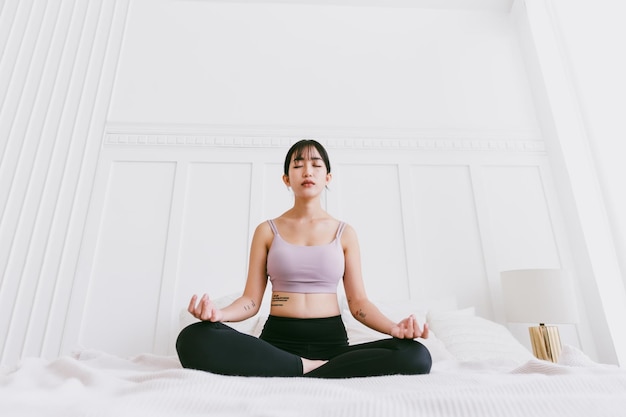 Sportliche asiatische Frau, die Yoga auf einem weißen Bett praktiziert und dabei Ardha Padmasana-Übungen macht, die in halber Lotus-Pose meditieren und zu Hause in Sportkleidung trainieren