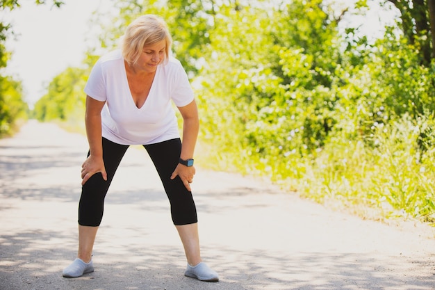 Foto sportliche blonde frau, die sich nach dem joggen im park morgens ausruht, eine intelligente uhr trägt und ihre hände auf den knien hält. foto in hoher qualität