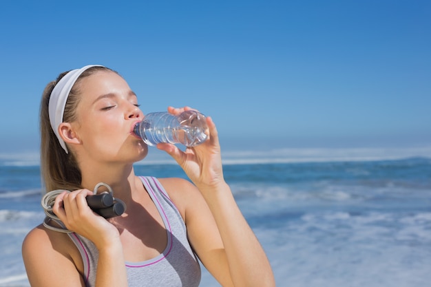 Sportliche Blondine, die auf dem Strand mit Flasche und Springseil an einem sonnigen Tag steht