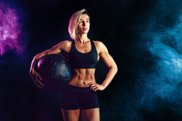 Sportliche Blondine in der modernen Sportkleidung, die mit Medizinball aufwirft. Foto der muskulösen Frau auf dunkler Wand mit Rauche. Kraft und Motivation.