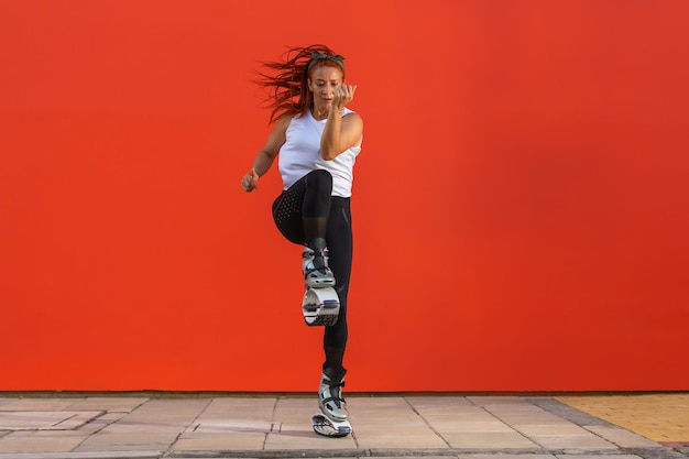 Sportliche Frau, die eine Fitnessroutine mit Kangoo Jumps-Stiefeln auf orangem Hintergrund macht. Gesundes Lebens-, Aktivitäts- und Sportkonzept