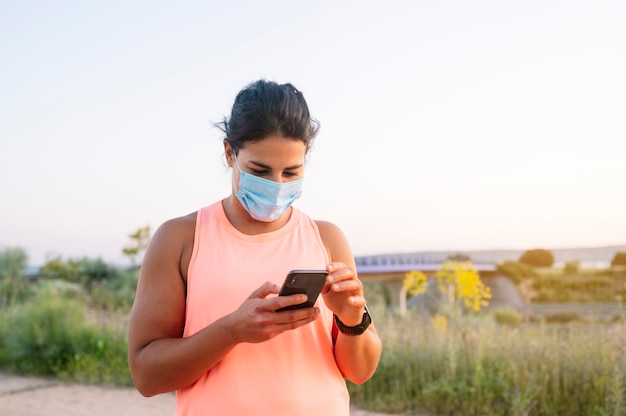 Sportliche Frau, die ihr Smartphone mit Gesichtsmaske im Freien verwendet