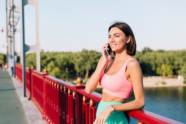 Sportliche Frau in passender Sportkleidung bei Sonnenuntergang an der modernen Brücke mit Blick auf den Fluss glückliches positives Lächeln mit Handygespräch mit Gespräch