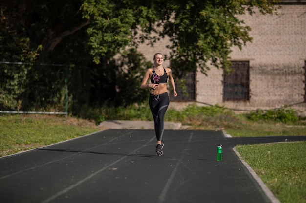 Sportliche Frau mit glücklichem Gesichtsausdruck im Rücken-T-Shirt und Leggings im Stadion mit einer Flasche Wasser