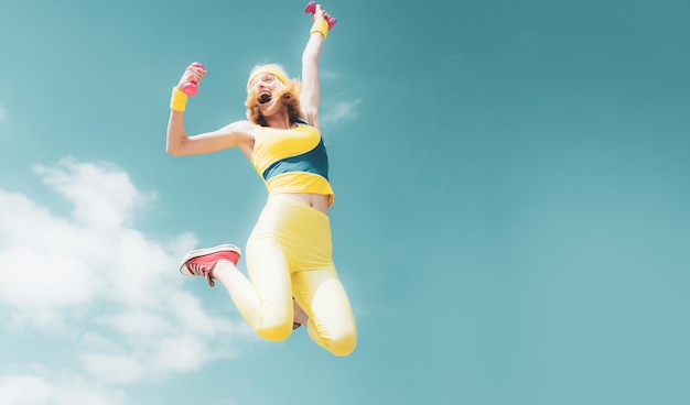 Sportliche Frau springt mit Hanteln lustige Frau in Sportkleidung auf Himmelshintergrund dynamische Bewegung spo
