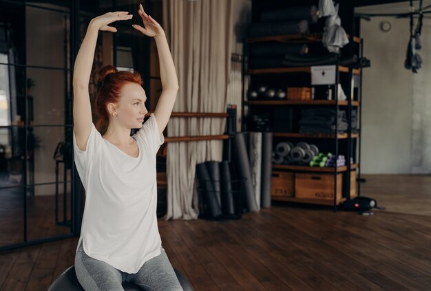 Sportliche, grazile junge Frau mit roten Haaren sitzt auf einem großen silbernen Pilates-Ball mit über den Kopf gehobenen Händen, die sich vor dem Training im Fitnessstudio aufwärmen. Konzept des gesunden Lebensstils und des Sports indoor