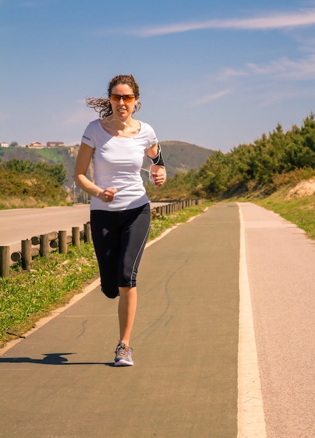 Sportliche junge Frau mit Kopfhörern, die Musik von ihrem Smartphone hört, während sie auf einer Landebahn im Freien läuft. Modernes gesundes Lebensstilkonzept.