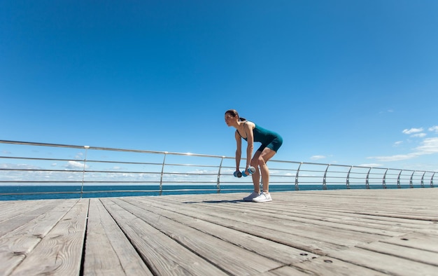 Sportliche kaukasische Frau trainiert mit Hanteln auf dem Damm an einem hellen sonnigen Tag mit blauem Himmel und Wolken Gesundes Lebensstil-Konzept Weitwinkel