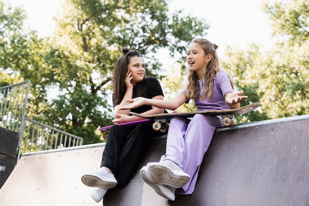 Sportliche Kinder Mädchen mit Skateboard und Penny Boards sitzen und plaudern auf der Sportrampe auf dem Spielplatz Freunde Kommunikation und Freundschaft Sport extremer Lebensstil